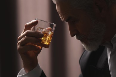 Photo of Senior man holding glass of whiskey with ice cubes on brown background, closeup