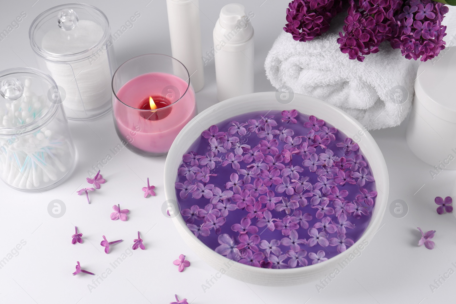 Photo of Bowl of water with lilac flowers, spa products and burning candle on white table, above view
