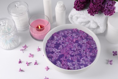 Photo of Bowl of water with lilac flowers, spa products and burning candle on white table, above view