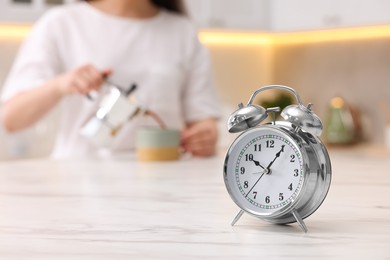 Photo of Alarm clock on white marble table. Woman pouring coffee from jezve into cup in kitchen, selective focus