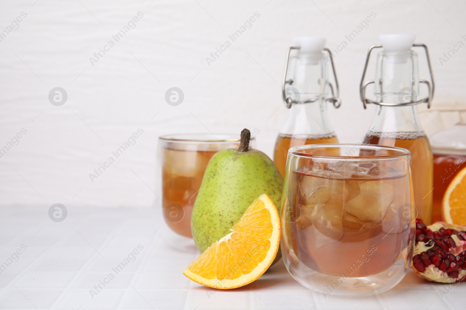 Photo of Tasty kombucha with ice cubes and fresh fruits on white tiled table, space for text