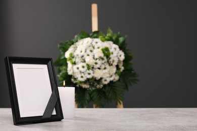 Photo frame with black ribbon, burning candle on light table and wreath of flowers near grey wall indoors, space for text. Funeral attributes