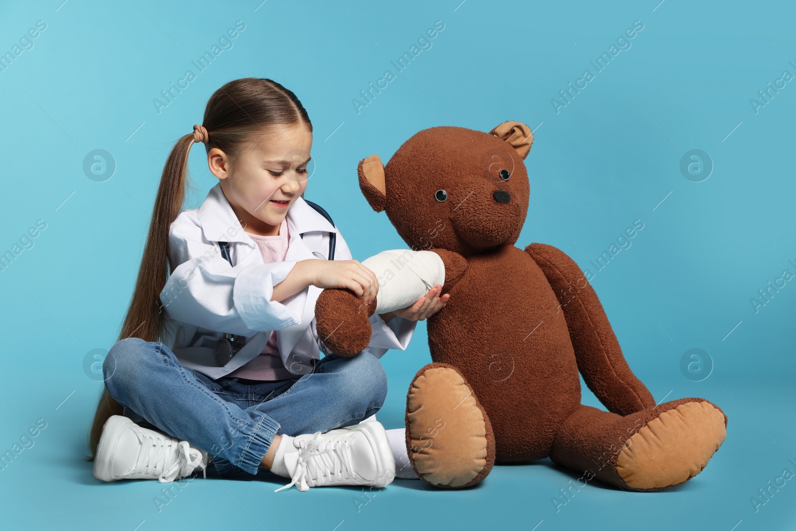 Photo of Little girl in medical uniform and toy bear with bandage on light blue background