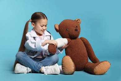 Photo of Little girl in medical uniform and toy bear with bandage on light blue background