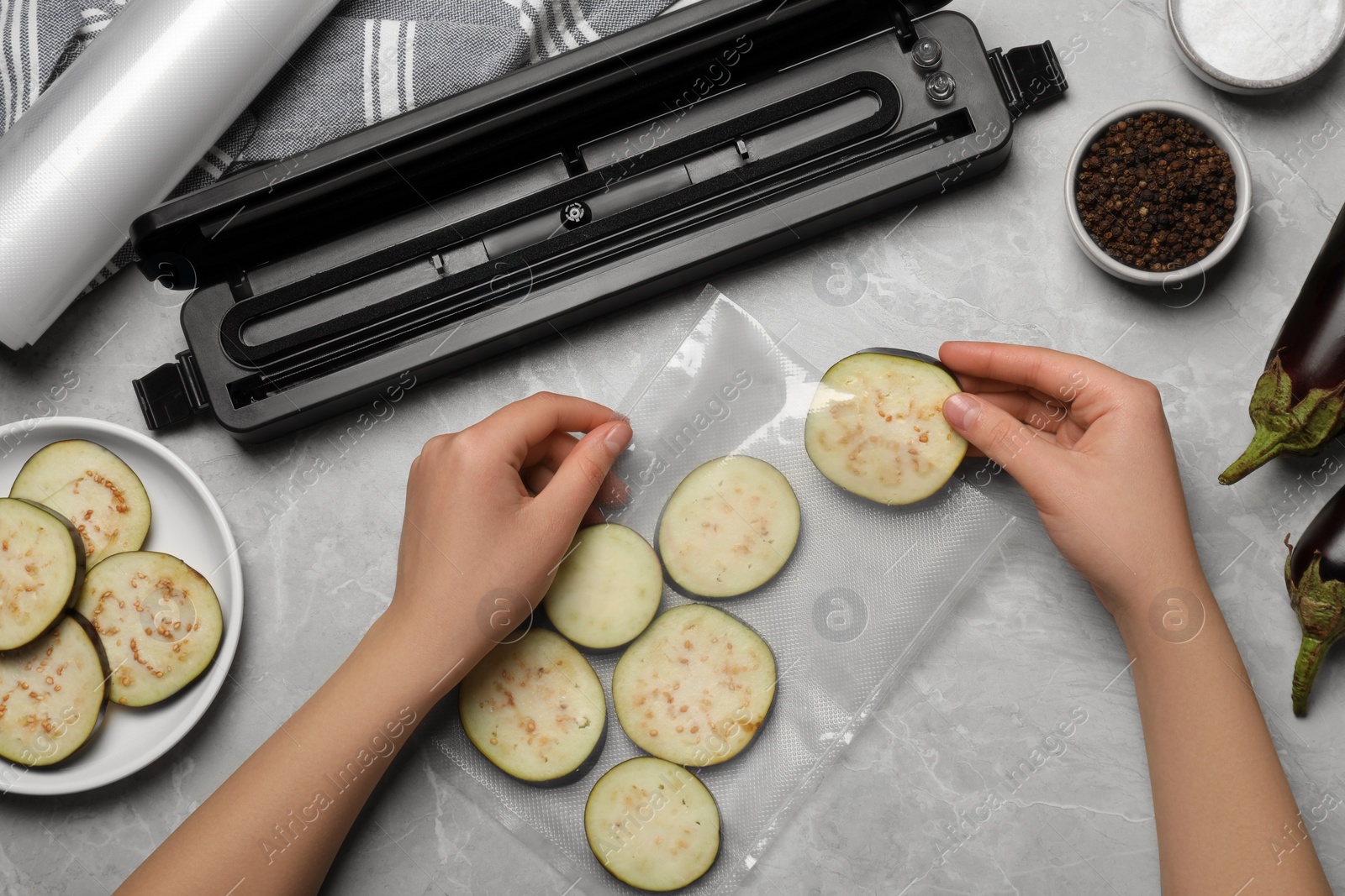 Photo of Woman packing cut eggplant into plastic bag using vacuum sealer on light grey marble table, closeup