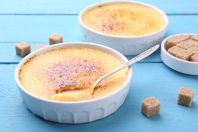 Photo of Delicious creme brulee in bowls, sugar cubes and spoon on light blue wooden table, closeup