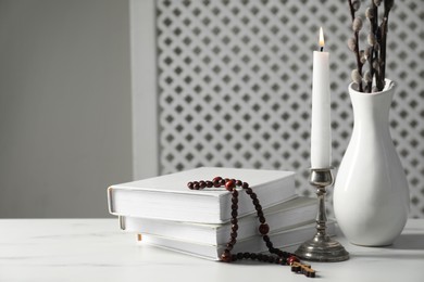 Books, burning candle, rosary beads and vase of willow branches on white marble table, closeup