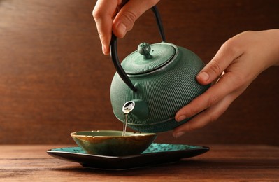 Photo of Woman pouring freshly brewed tea from teapot into cup at wooden table, closeup. Traditional ceremony
