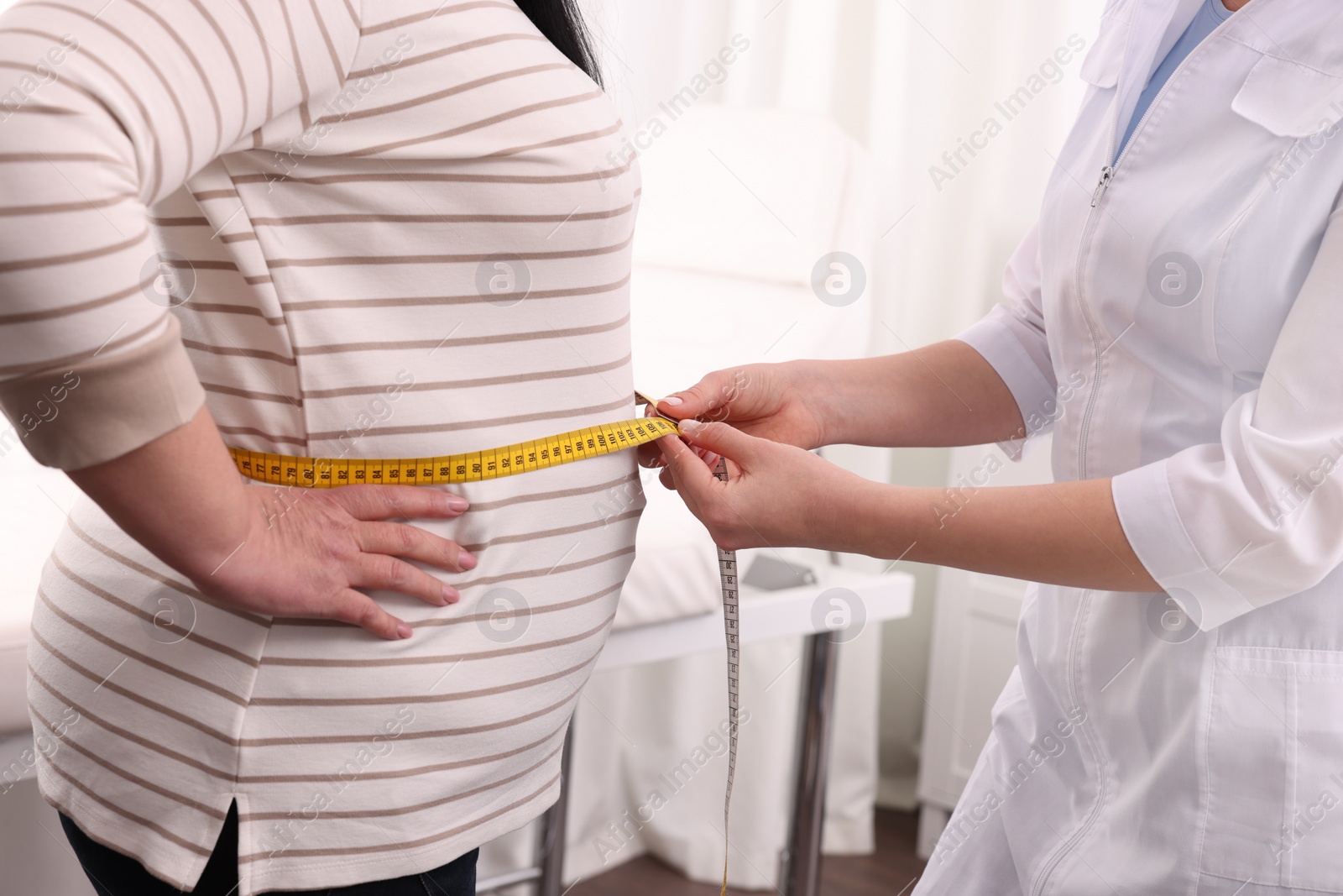 Photo of Nutritionist measuring overweight woman's waist with tape in clinic, closeup