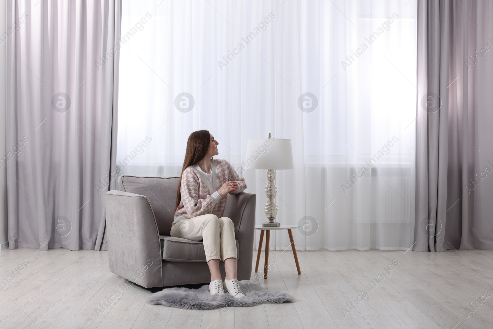 Photo of Woman sitting on armchair near lamp and window with stylish curtains at home. Space for text