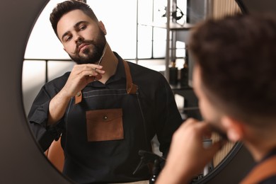 Handsome young man trimming beard with scissors near mirror indoors