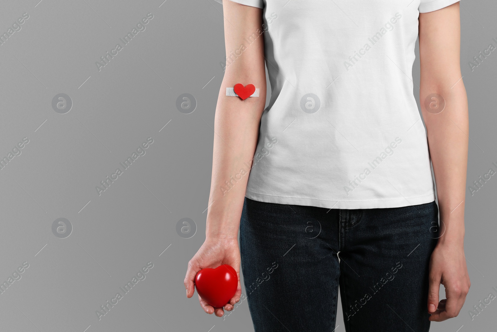 Photo of Blood donation concept. Woman with adhesive plaster on arm holding red heart against grey background, closeup. Space for text