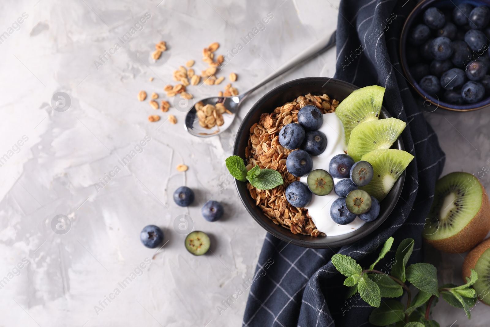 Photo of Tasty granola with yogurt, blueberries and kiwi in bowl on gray textured table, flat lay. Space for text