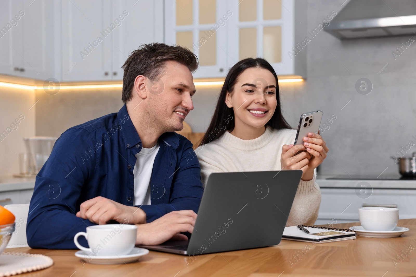 Photo of Happy couple with gadgets shopping online at wooden table in kitchen