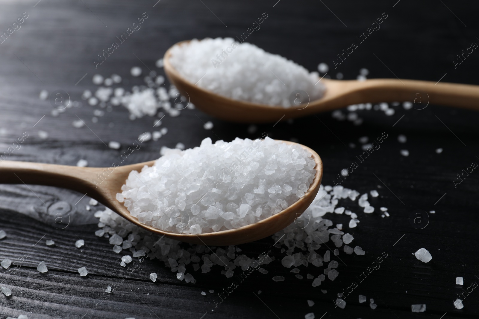 Photo of Organic salt in spoons on black wooden table, closeup