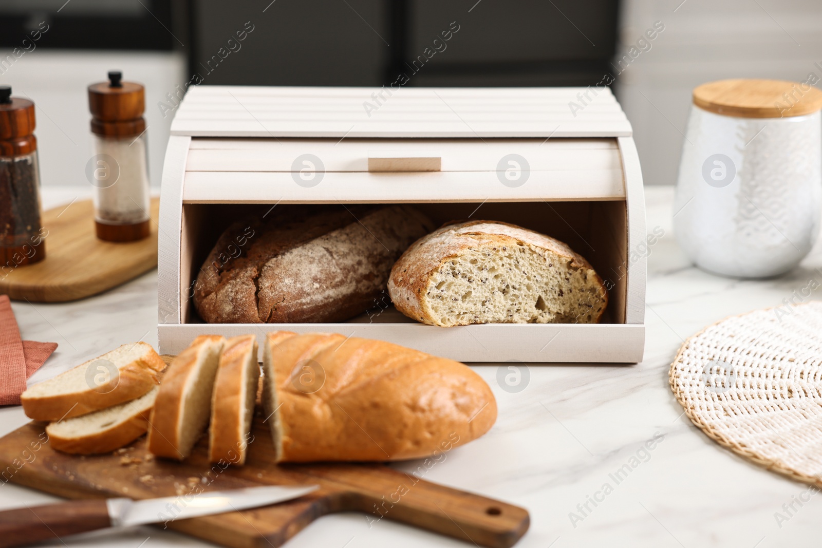 Photo of Wooden bread basket with freshly baked loaves and knife on white marble table in kitchen