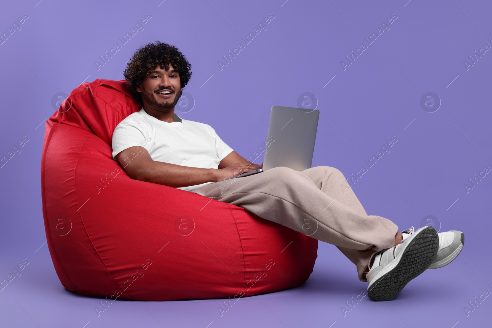 Photo of Smiling man with laptop sitting on beanbag chair against purple background