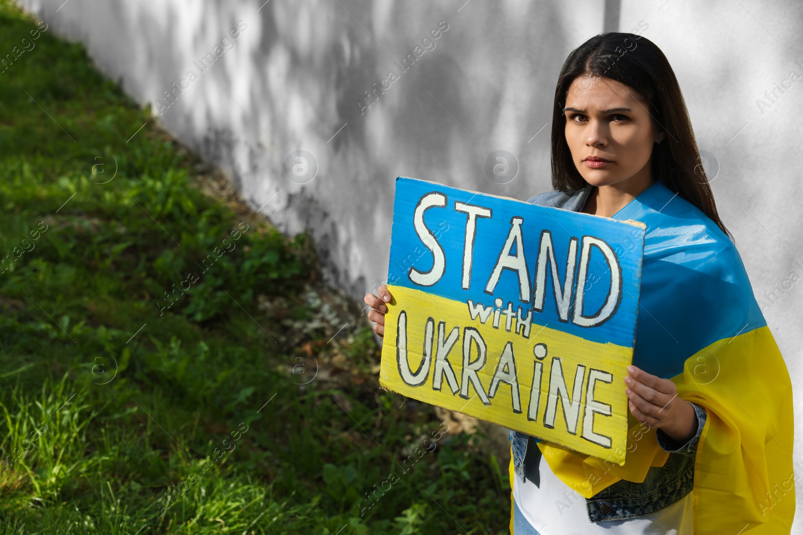 Photo of Sad woman holding poster in colors of national flag and words Stand with Ukraine outdoors. Space for text