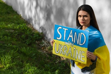 Photo of Sad woman holding poster in colors of national flag and words Stand with Ukraine outdoors. Space for text