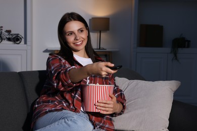 Happy woman holding popcorn bucket and changing TV channels with remote control at home in evening