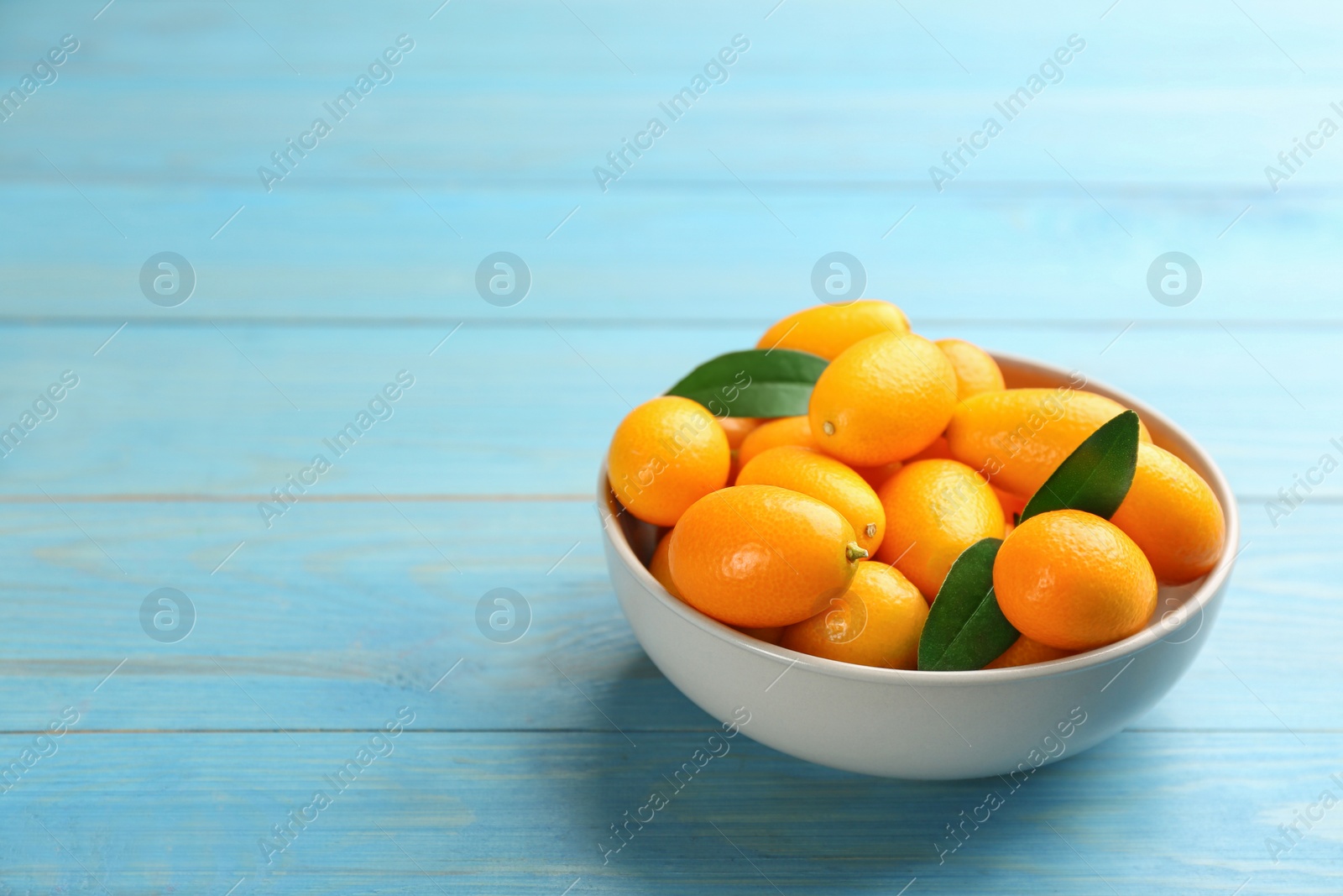 Photo of Fresh ripe kumquats in bowl on light blue wooden table, space for text