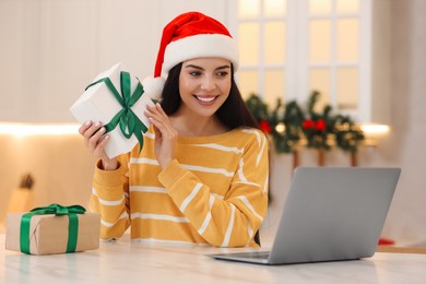 Photo of Celebrating Christmas online with exchanged by mail presents. Smiling woman in Santa hat with gift boxes during video call on laptop at home