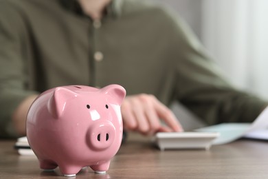 Financial savings. Man using calculator at wooden table, focus on piggy bank