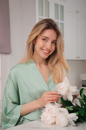 Photo of Pretty young woman in beautiful silk robe with flowers at kitchen table