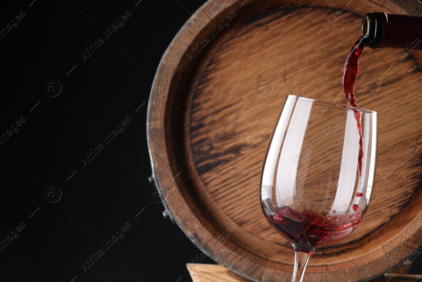 Photo of Pouring delicious wine into glass near wooden barrel against black background, closeup. Space for text