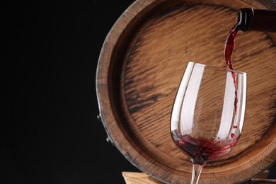 Pouring delicious wine into glass near wooden barrel against black background, closeup. Space for text