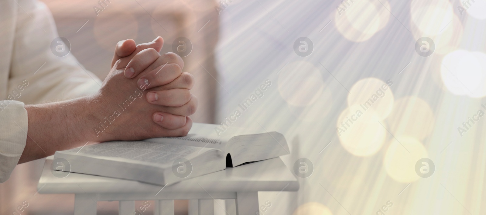 Image of Religion. Christian man praying over Bible at table, closeup. Bokeh effect. Banner design