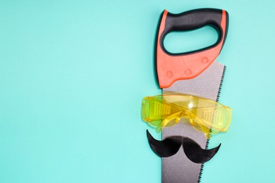 Photo of Man's face made of artificial mustache, safety glasses and hand saw on light blue background, top view. Space for text