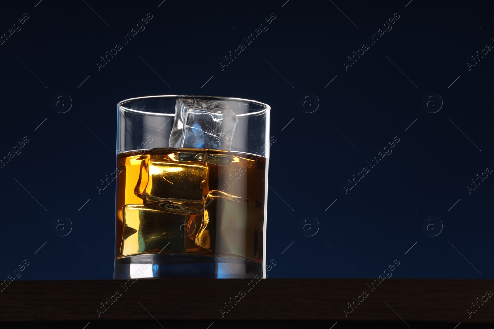 Photo of Whiskey with ice cubes in glass on table against dark blue background. Space for text