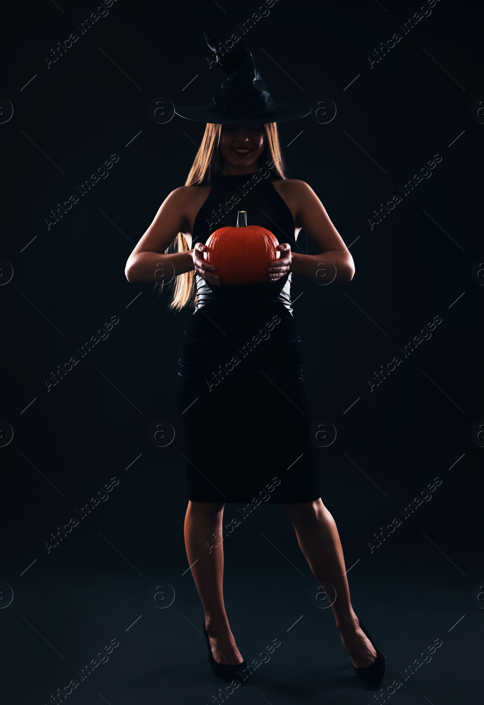 Photo of Young woman wearing witch costume with pumpkin on black background. Halloween party