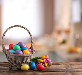 Image of Colorful Easter eggs in wicker basket and tulips on wooden table indoors, space for text 