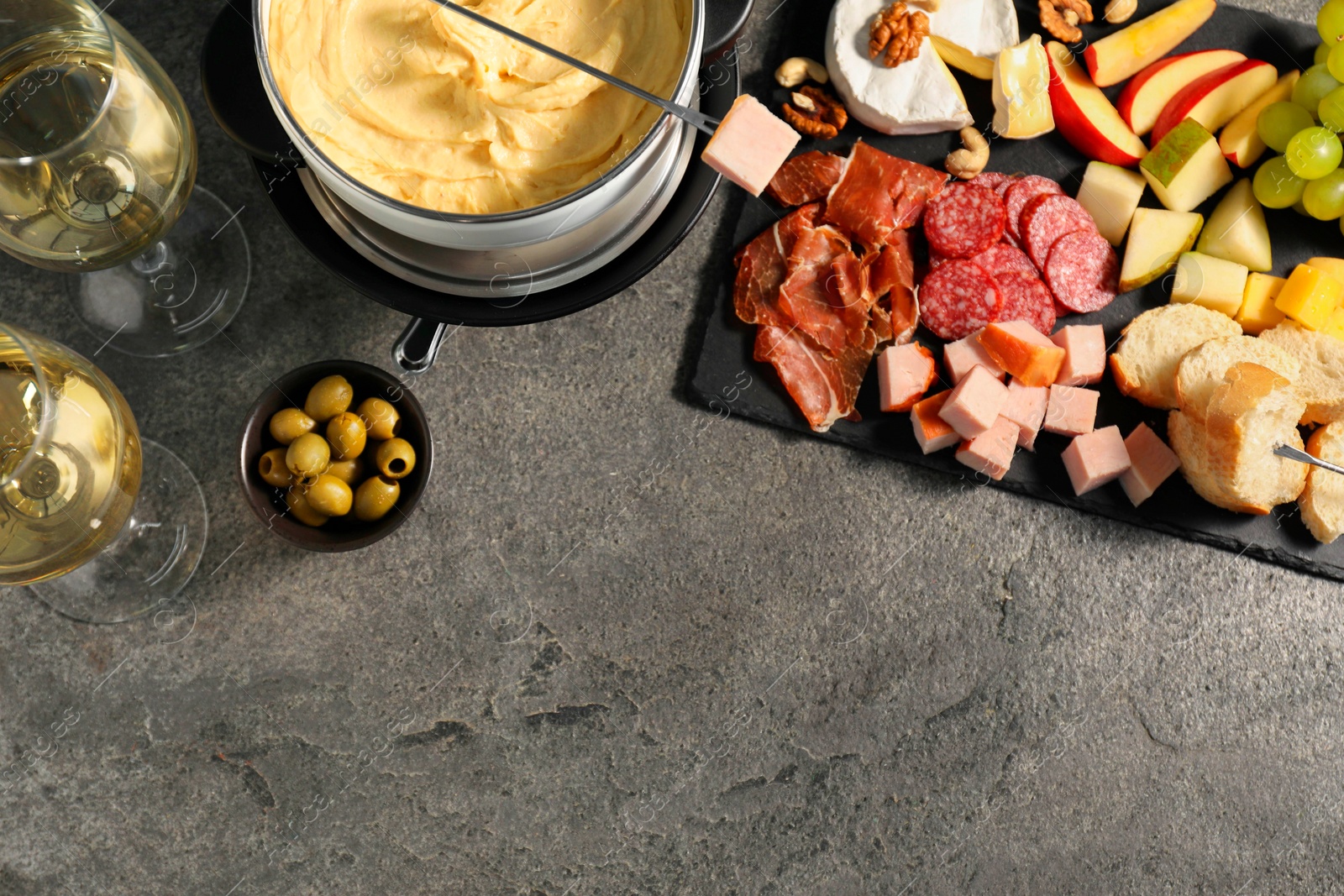 Photo of Fork with piece of ham, fondue pot with melted cheese, wine and snacks on grey textured table, flat lay. Space for text