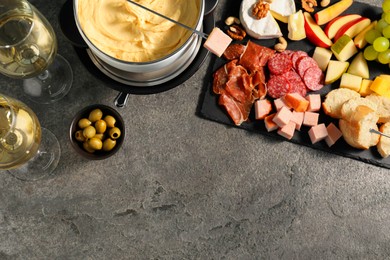 Photo of Fork with piece of ham, fondue pot with melted cheese, wine and snacks on grey textured table, flat lay. Space for text