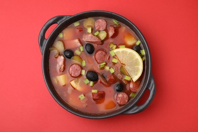 Photo of Meat solyanka soup with thin dry smoked sausages in bowl on red background, top view