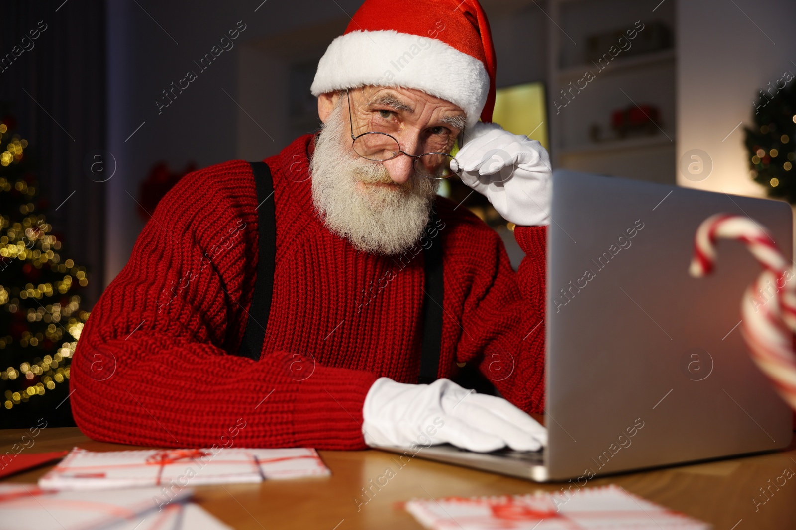 Photo of Santa Claus using laptop at his workplace in room decorated for Christmas