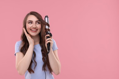 Beautiful young woman using curling hair iron on pink background, space for text
