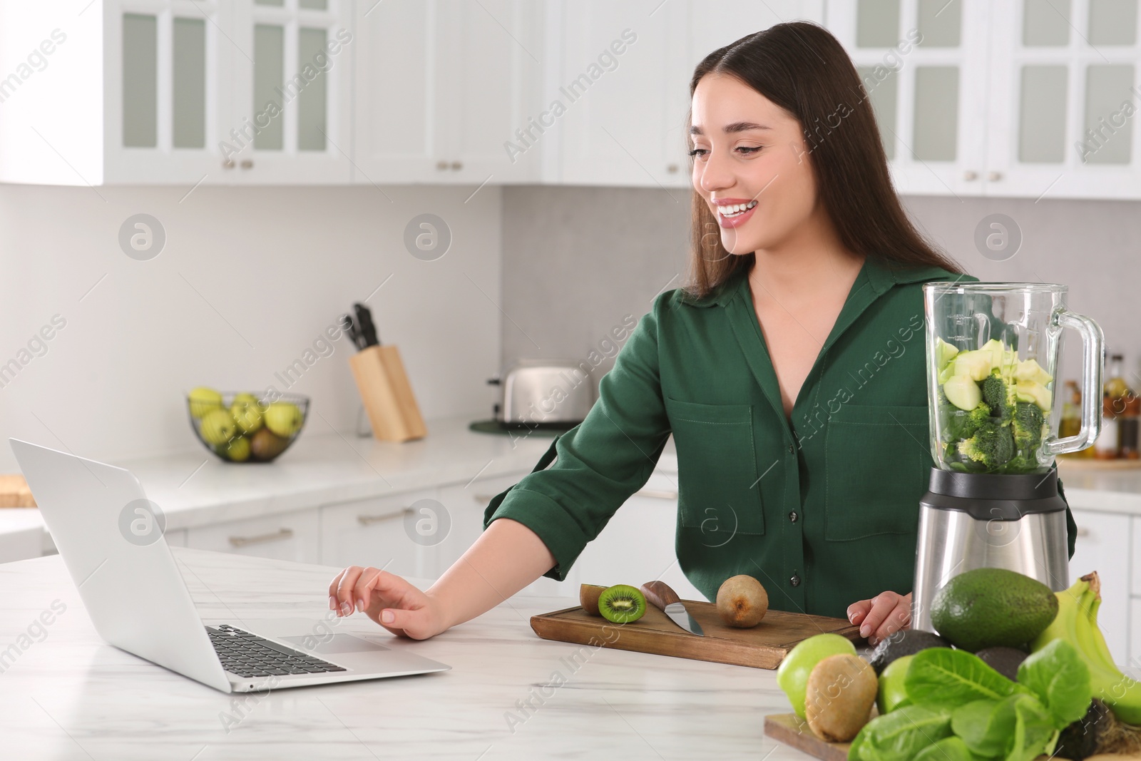 Photo of Young woman using laptop while preparing smoothie at white table in kitchen