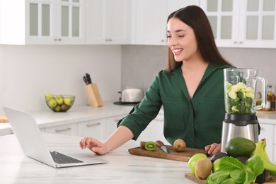 Young woman using laptop while preparing smoothie at white table in kitchen