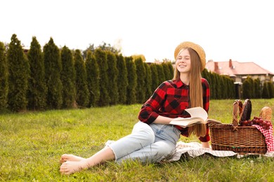 Happy girl with picnic basket reading book on green grass in park