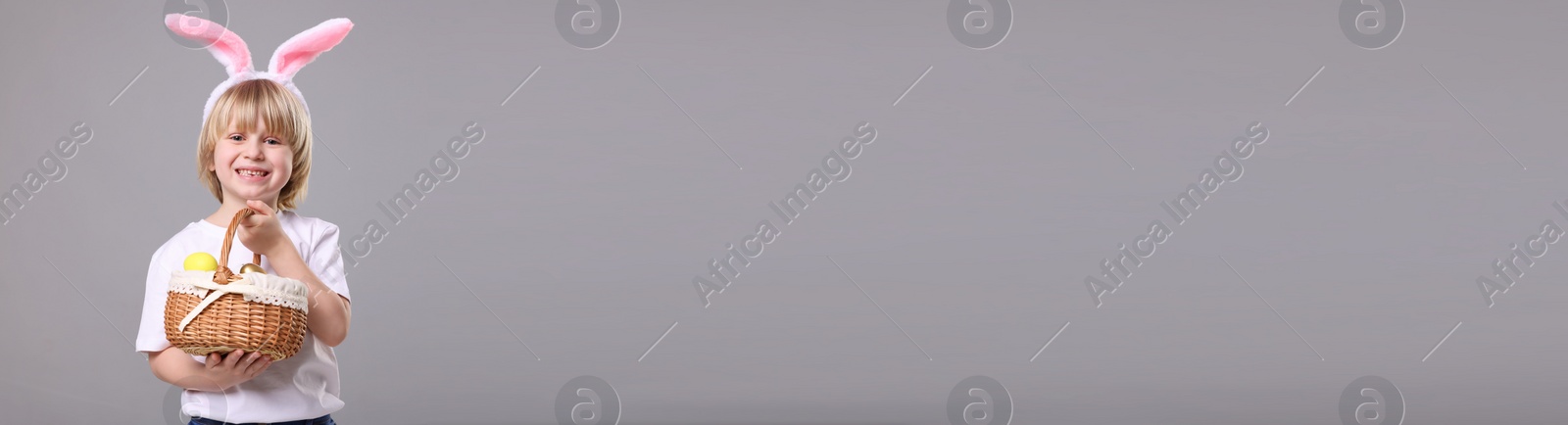 Image of Happy boy with bunny ears holding basket full of Easter eggs on light grey background, space for text. Banner design