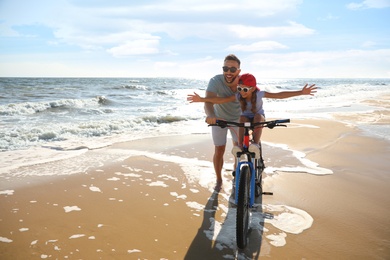 Happy father teaching daughter to ride bicycle on sandy beach near sea