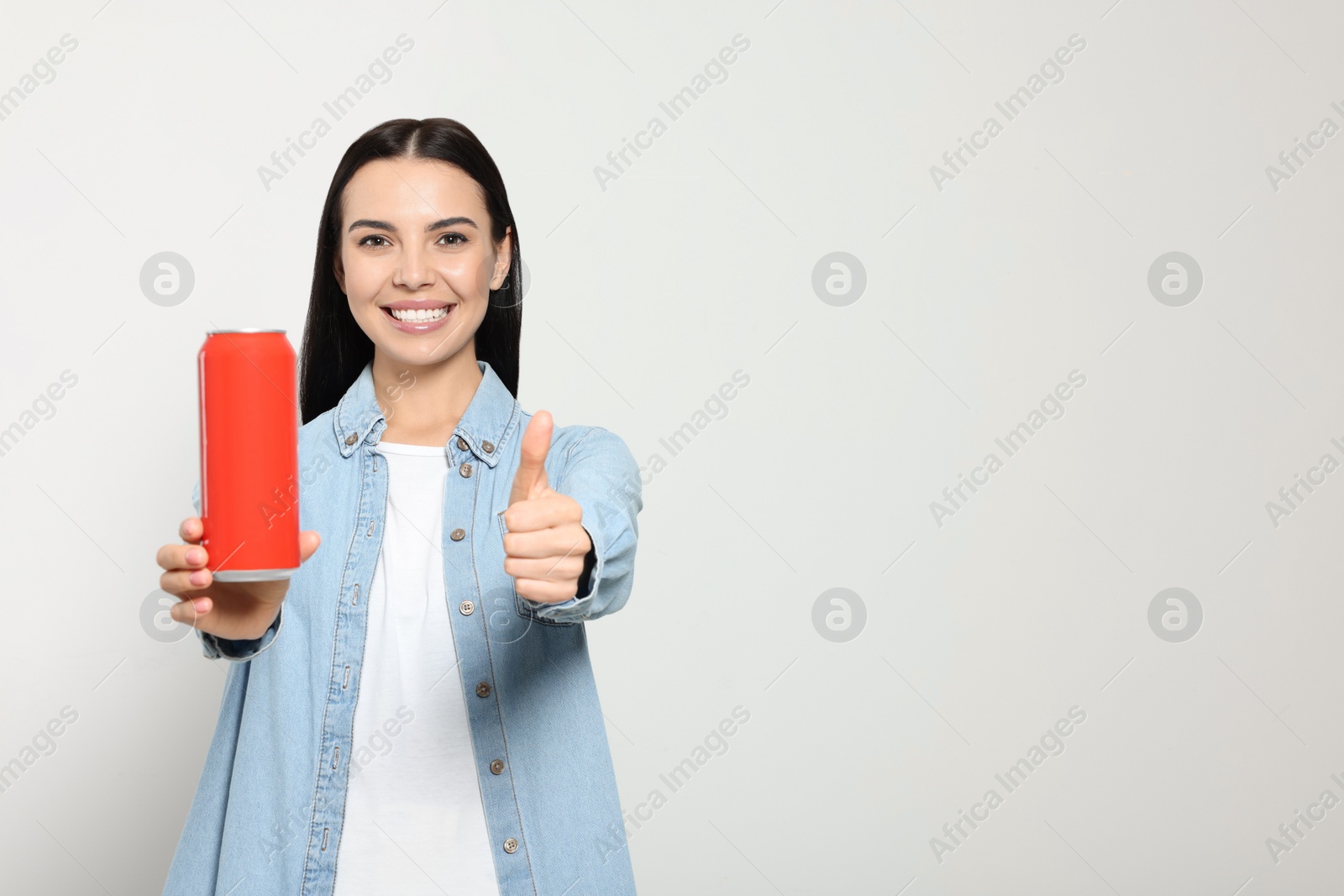 Photo of Beautiful happy woman holding red beverage can and showing thumbs up on light grey background. Space for text