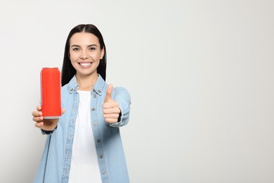 Beautiful happy woman holding red beverage can and showing thumbs up on light grey background. Space for text