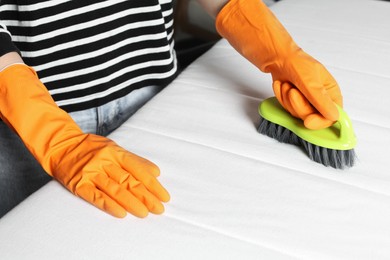 Photo of Woman in orange gloves cleaning white mattress with brush indoors, closeup