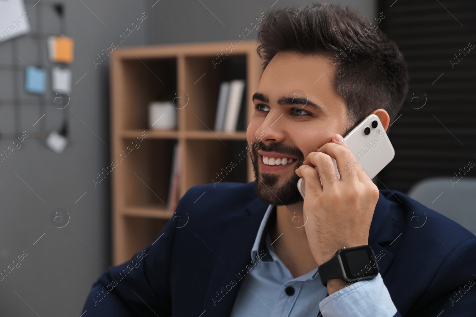 Photo of Happy young businessman wearing smart watch and talking on smartphone in office