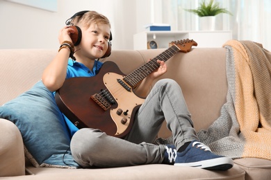 Photo of Cute little boy with headphones playing guitar on sofa in room